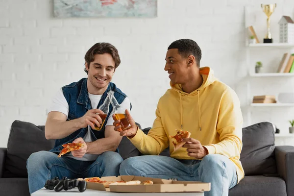 KYIV, UKRAINE - MARCH 22, 2021: interracial hipsters clinking with beer bottles on couch in living room — Stock Photo