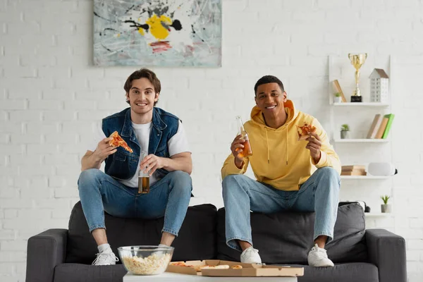 Felices amigos interracial disfrutando de rebanadas de pizza, palomitas de maíz y cerveza en el sofá en la sala de estar moderna - foto de stock