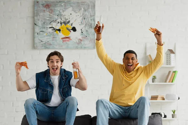 Interracial friends enjoying pizza slices, and beer with win gestures on couch in modern living room — Stock Photo