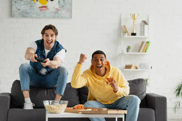 KYIV, UCRANIA - 22 de marzo de 2021: amigos interracial jugando videojuegos con joystick y disfrutando de la pizza en el sofá en la moderna sala de estar - foto de stock