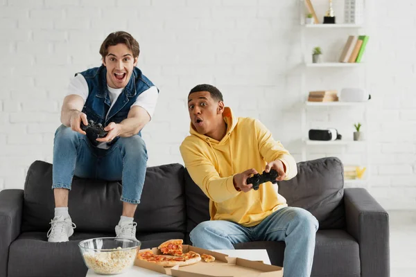 KYIV, UCRANIA - 22 de marzo de 2021: amigos interracial jugando emocionalmente a videojuegos con joysticks y disfrutando de la pizza en el sofá en la moderna sala de estar - foto de stock