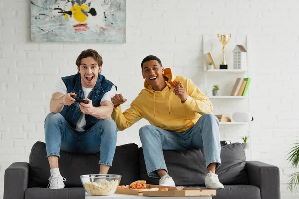 KYIV, UKRAINE - MARCH 22, 2021: interracial friends playing video game with joystick and enjoying pizza and popcorn on couch in modern living room — Stock Photo