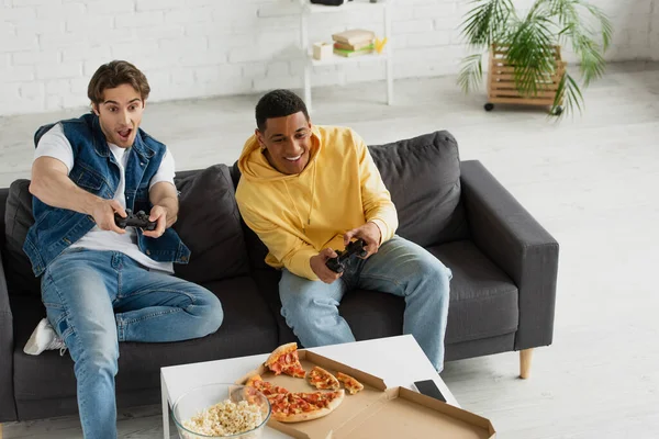 KYIV, UKRAINE - 22 MARS 2021 : vue en angle élevé d'amis interraciaux jouant émotionnellement à un jeu vidéo avec des joysticks et dégustant une pizza sur le canapé dans le salon moderne — Photo de stock