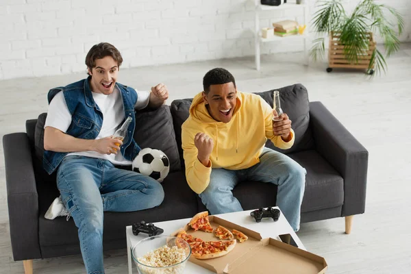 QUIIV, UCRÂNIA - MARÇO 22, 2021: visão de alto ângulo dos fãs de futebol interracial emocionalmente assistindo jogo juntos no sofá na sala de estar — Fotografia de Stock