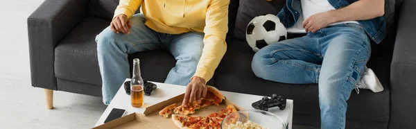 KYIV, UCRANIA - 22 DE MARZO DE 2021: vista parcial de los hombres en jeans sentados en el sofá cerca de la mesa con pizza, botella de cerveza, palomitas de maíz, joysticks y pelota, pancarta - foto de stock