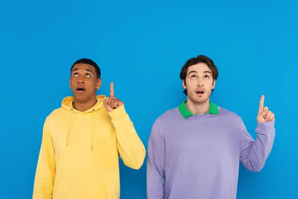 Amigos interracial mirando hacia arriba y mostrando la idea signo con las manos aisladas en azul - foto de stock