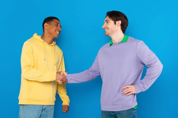 Sonrientes amigos interracial estrechando las manos aisladas en azul - foto de stock