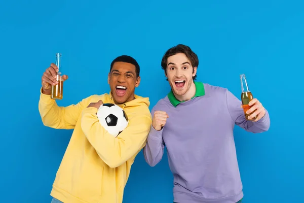 Sonrientes amigos interracial sosteniendo botellas de cerveza y fútbol aislados en azul - foto de stock