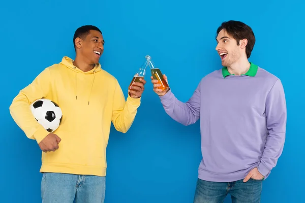 Улыбающиеся межрасовые друзья, держащие футбол и тосты с бутылками пива, изолированными на голубом — стоковое фото
