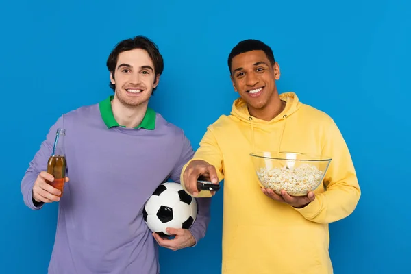 Interracial football fans holding tv emote controller, bottle of beer and popcorn isolated on blue — Stock Photo