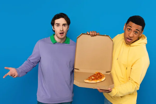 Hipster interrazziale in possesso di scatola con fetta di pizza isolato su blu — Foto stock