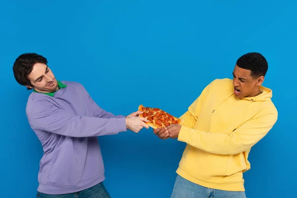 Interrassische Freunde teilen emotional eine Pizza-Scheibe isoliert auf blauem Grund — Stockfoto