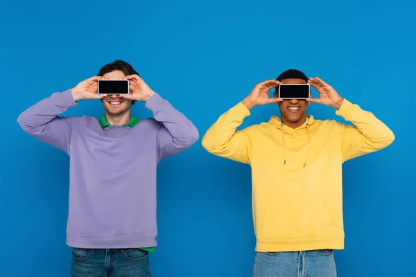 Interracial hipsters covering eyes with blank screen smartphones isolated on blue — Stock Photo