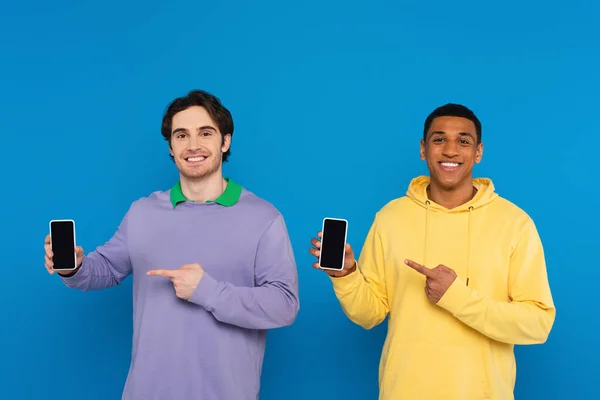 Amigos inter-raciais apontando com os dedos em smartphones de tela em branco com telas em branco isolados em azul — Fotografia de Stock