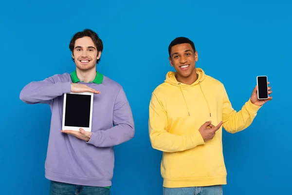 Amigos interracial feliz mostrando dispositivos digitales con pantalla en blanco aislado en azul - foto de stock