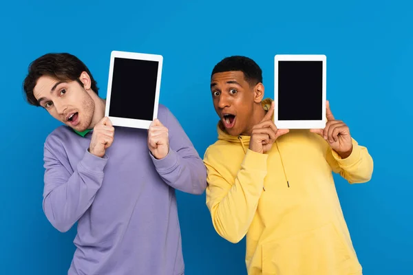 Amigos interraciales con caras alegres sosteniendo dispositivos digitales cerca de caras con la pantalla en blanco aislada en azul - foto de stock