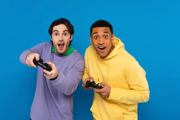 KYIV, UKRAINE - MARCH 22, 2021: friends with joyful faces holding joysticks and looking at camera isolated on blue — Stock Photo