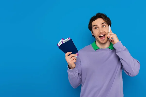 Heureux jeune homme tenant des passeports avec des billets d'avion dans les mains et parlant sur téléphone cellulaire isolé sur bleu — Photo de stock