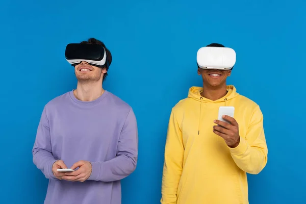 Hipsters inter-raciais em fones de ouvido de realidade virtual e smartphones isolados em azul — Fotografia de Stock