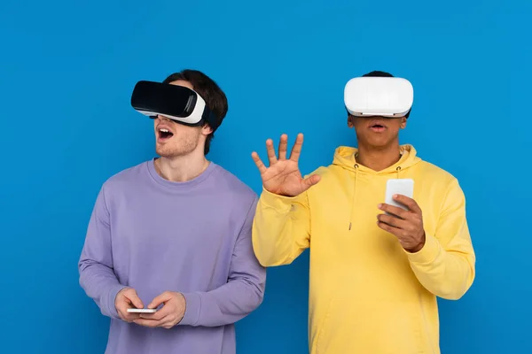 Animado interracial hipsters jogar vídeo game com virtual realidade headsets isolado no azul — Fotografia de Stock