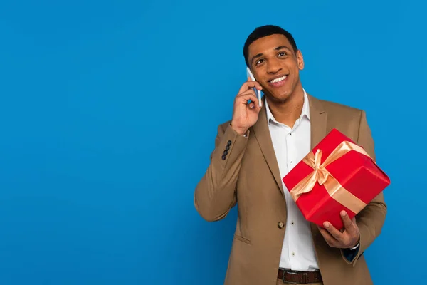 Joven afroamericano hombre en traje de moda sosteniendo caja de regalo y hablando por teléfono celular aislado en azul - foto de stock