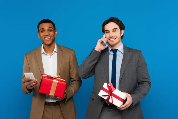 Sorrindo jovens homens inter-raciais em ternos elegantes segurando caixas de presente com fitas vermelhas e douradas e usando smartphones isolados em azul — Fotografia de Stock
