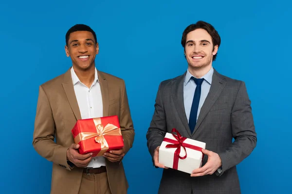 Sonrientes jóvenes hombres interracial en trajes de moda sosteniendo cajas de regalo con cintas rojas y doradas aisladas en azul - foto de stock