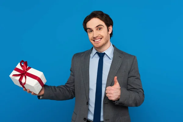 Sorrindo jovem em blazer segurando caixa de presente e mostrando polegares para cima gesto isolado no azul — Fotografia de Stock