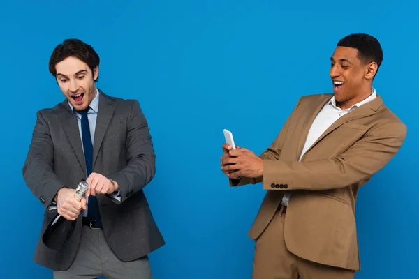 Smiling african american man filming with smartphone how his friend opening bottle of champagne isolated on blue — Stock Photo