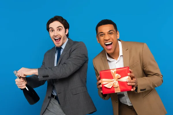 Joyeux interracial amis en costumes s'amuser avec boîte cadeau et bouteille de champagne isolé sur bleu — Photo de stock