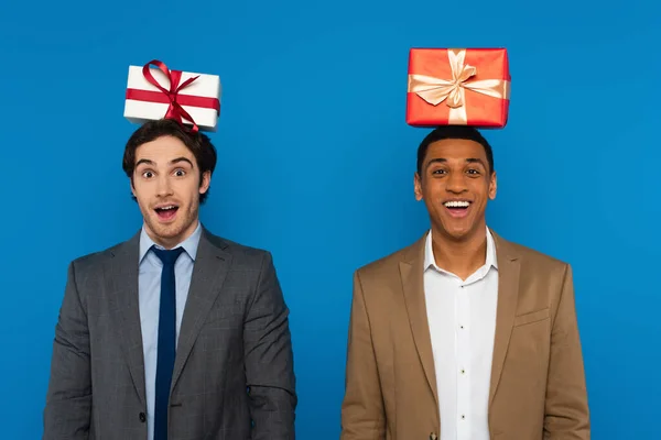 Joyful interracial friends in suits smiling with gift boxes on heads isolated on blue — Stock Photo