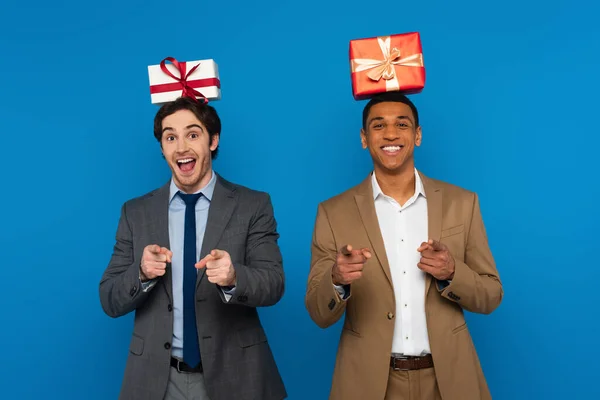 Alegres amigos interracial en trajes sosteniendo cajas de regalo en cabezas aisladas en azul - foto de stock