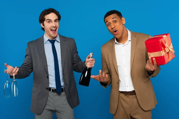 Joyful interracial friends in suits holding champagne bottle, glasses and gift box isolated on blue — Stock Photo