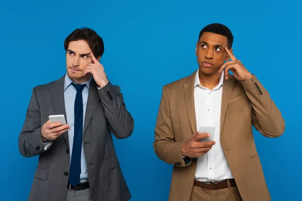 Задумчивые межрасовые мужчины с вдумчивым жестом, держащие смартфоны в руках изолированные на голубом — стоковое фото