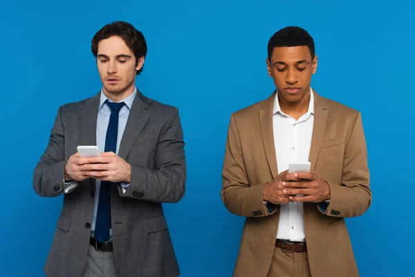 Interracial men in suits watching at smartphones in hands isolated on blue — Stock Photo