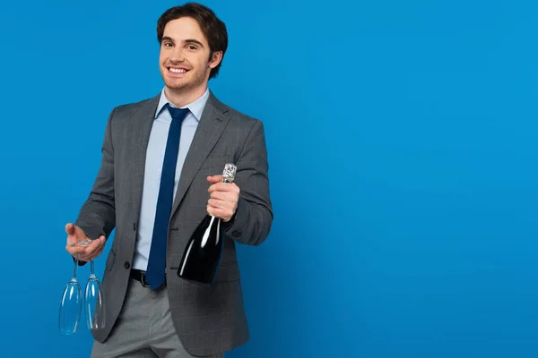 Joven sonriente en traje elegante sosteniendo botella de champán y copas aisladas en azul, pancarta - foto de stock