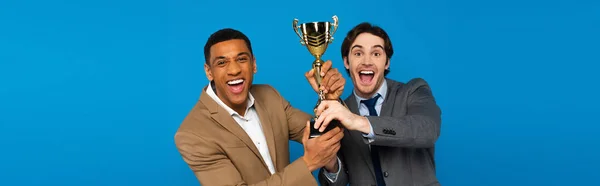 Interracial friends in suits celebrating success with prize cup in hands isolated on blue, banner — Stock Photo