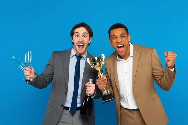 Amigos interracial en trajes celebrando el éxito con copa de triunfo, champán y copas en manos aisladas en azul - foto de stock