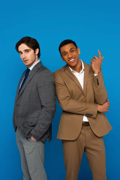Elegantes hombres interracial en trajes de pie con confianza aislado en azul - foto de stock
