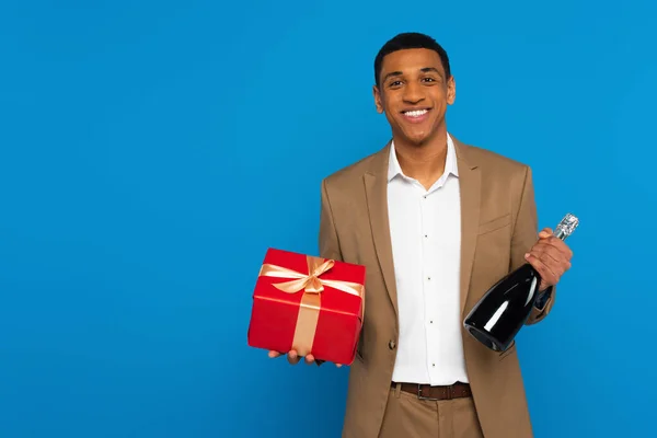 Sonriente hombre afroamericano en traje elegante sosteniendo botella de champán y caja de regalo aislado en azul - foto de stock
