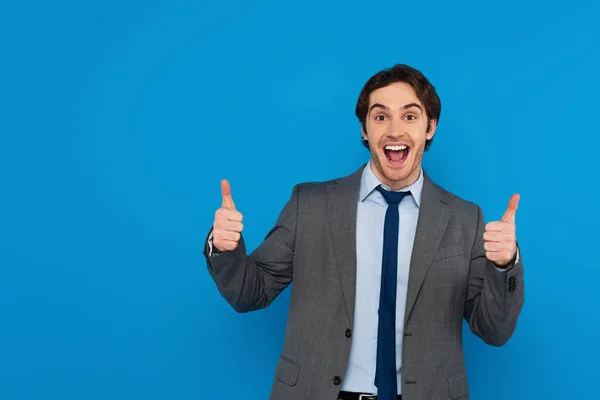 Счастливый улыбчивый мужчина в костюме, показывающий большим пальцем вверх жест на синем фоне — стоковое фото