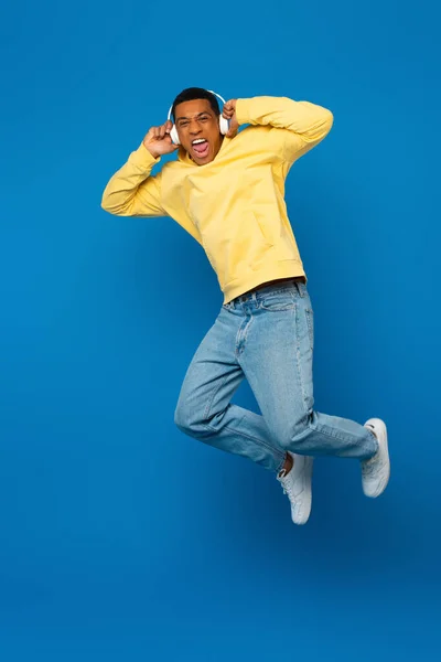 Excited african american man jumping in headphones on blue background — Stock Photo