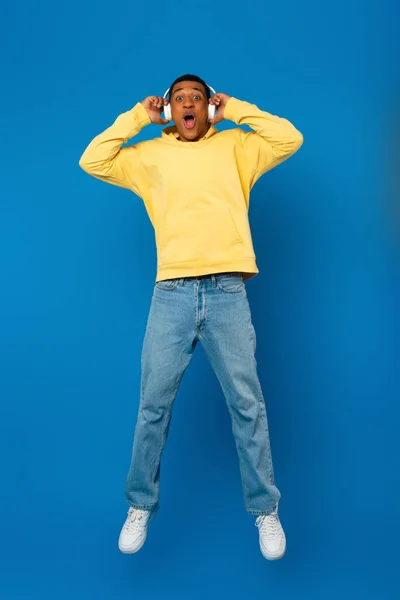 Hombre afroamericano feliz saltando en auriculares sobre fondo azul - foto de stock