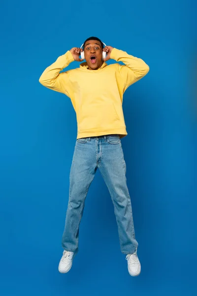 Happy african american man jumping in headphones on blue background — Stock Photo