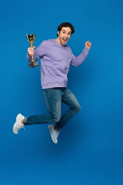 Feliz hombre sonriente en sudadera violeta saltando con copa de trofeo sobre fondo azul - foto de stock