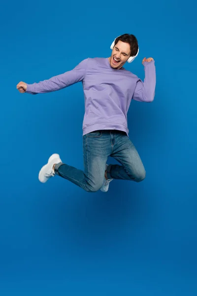 Feliz hombre sonriente en sudadera violeta disfrutando de la música y saltando en auriculares sobre fondo azul - foto de stock