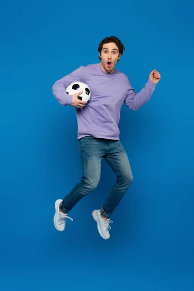 Glücklich lächelnder Mann im violetten Sweatshirt, der mit Fußball auf blauem Hintergrund springt — Stockfoto