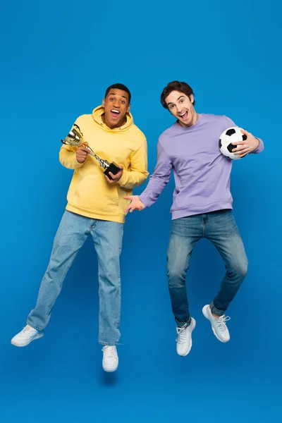 Happy african american man with friend levitating and holding football and cup on blue background — Stock Photo