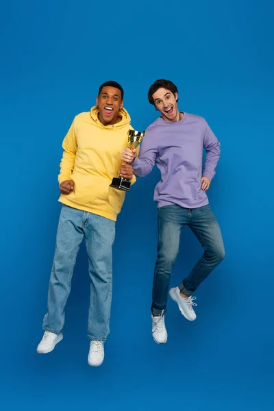 Happy african american man with friend levitating and holding triumph cup  in hands on blue background — Stock Photo