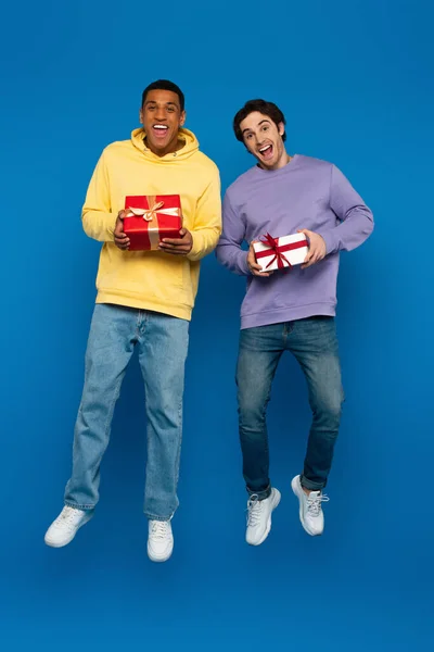 Hombre afroamericano feliz con amigo levitando con cajas de regalo en las manos sobre fondo azul - foto de stock